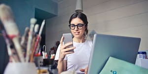 woman in white t shirt holding smartphone in front of laptop